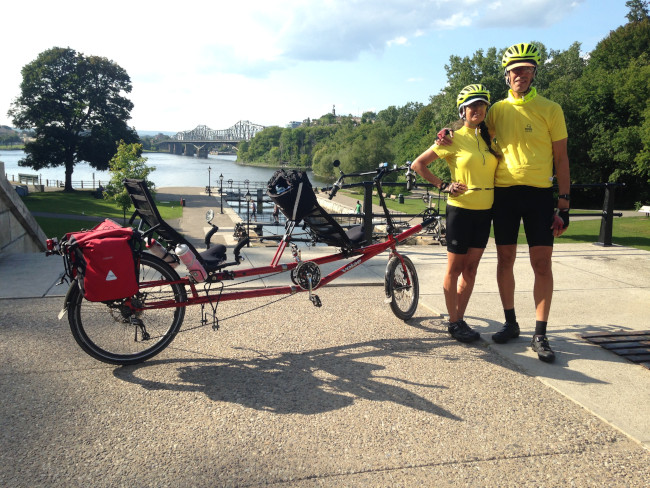 Couple standing by their tandem bike beside the 
        Rideau Canal with a view of the Ottawa River